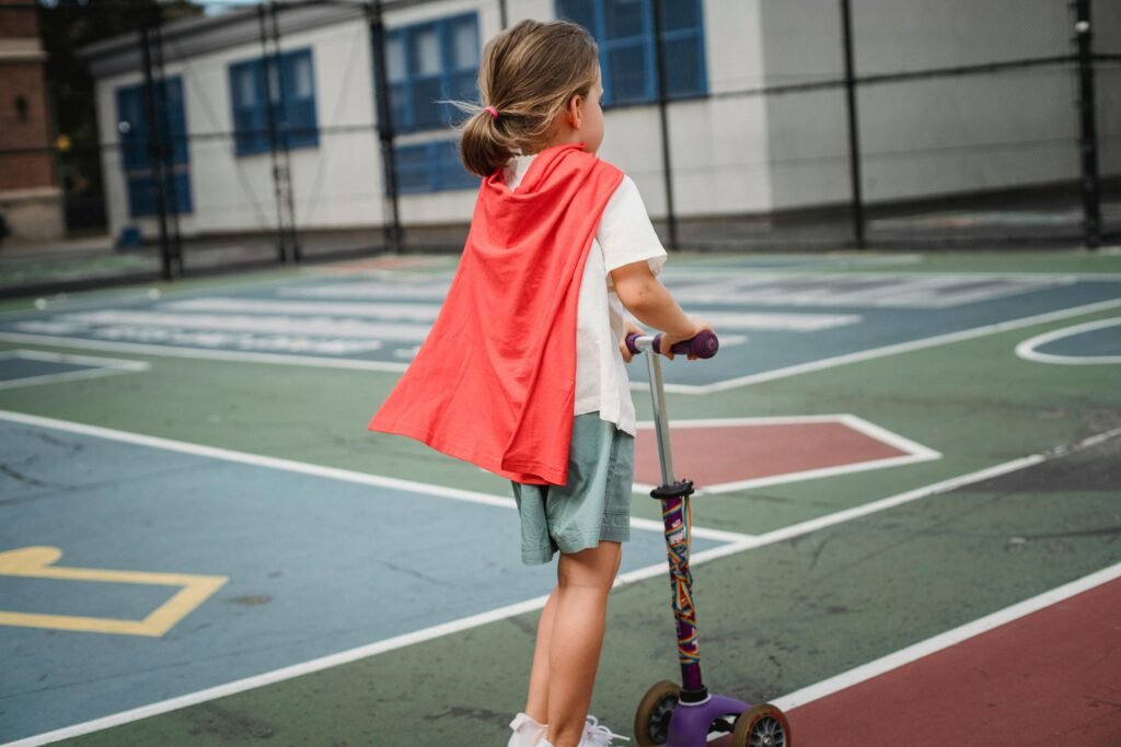 Young girl with cape on a scooter at a playground, back view.